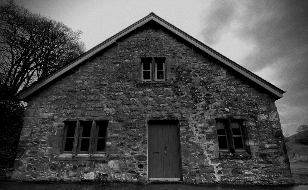 Black and white photograph of an old stone cottage beneath a cloudy sky, its dark windows and weathered door adding to the eerie atmosphere. The upper window appears to show the ghostly face of a little girl, a haunting nod to The Custodian’s Cottage, a short story from The Mereland Chronicles universe by Paul J. Scribbans.