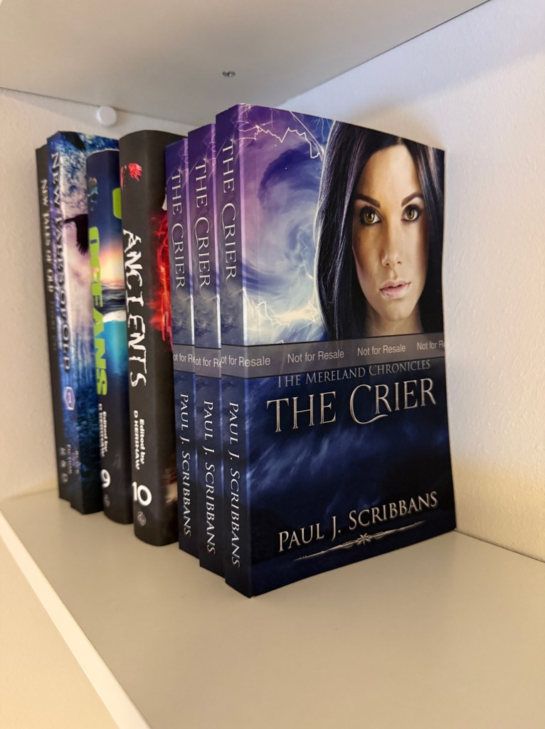 Proof copy of The Mereland Chronicles: The Crier by Paul J. Scribbans standing upright on a white bookshelf beside anthologies featuring his contributions with other authors.