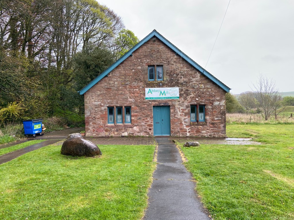 Abbey Mill Café building near Furness Abbey, a restored stone mill with blue-painted door and windows, set on grass with a paved path leading to the entrance.